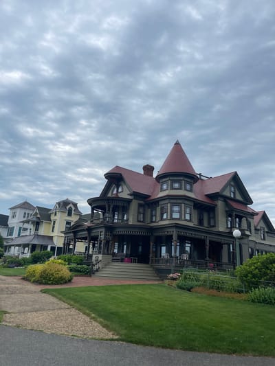 This media file shows a row of Victorian-style houses in Oak Bluffs, United States of America, under a cloudy sky. The largest house in the foreground is a dark green structure with a prominent conical red roof and intricate dark trim. It features a wide porch with decorative railings and columns. Above the windows of the conical turret, there is visible text in Latin: "ILLIUS GLORIQSVS LATINE". Further down the street, other colorful houses, including a yellow one, are visible. A well-maintained green lawn borders the sidewalk and the houses. The overall scene suggests a residential street on an overcast day. There are no people or specific activities depicted, but the architecture and landscaping suggest a well-preserved historical neighborhood.