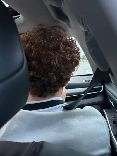 The image depicts the interior of a vehicle, viewed from behind a seated individual, presumably in Teaneck Township, United States. The central subject is the back of a person's head and upper torso. The individual has dense, tightly curled, reddish-brown hair. They are wearing a white, mesh-textured shirt with a black and white striped collar. A black seatbelt is diagonally secured across their right shoulder. To the left, part of a dark-colored car seat headrest is visible. The vehicle's light-colored headliner and a grab handle are visible at the top. Through the side window, an out-of-focus exterior scene is visible, characterized by diffused daylight and blurred colorful objects, possibly commercial signage or other vehicles, against a light-colored ground surface, consistent with an outdoor environment. Interior details like a silver trim strip, an air vent, and a black door panel with two window control buttons are also discernible on the right side of the frame. No direct interaction between individuals is depicted.
