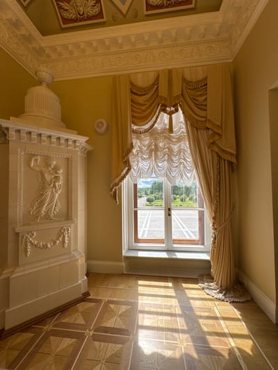 The image depicts an interior view of a room with ornate architectural details, located in Strelna, Russia. In the foreground, a parquet floor with a geometric pattern of light and dark wood is visible, with sunlight casting striped shadows across it. To the left, a tall, decorative fireplace or stove is featured, made of white stone or plaster with relief carvings of classical figures and floral motifs. The walls are painted a pale yellow. A large window dominates the right side of the room, draped with elaborate golden curtains adorned with tassels and fringe. Through the window, a view of a well-maintained park or garden with trees and pathways is visible. The ceiling features intricate stucco molding and painted decorative panels. A small white circular speaker is mounted on the wall near the window. The overall impression is one of grandeur and classical aesthetics.