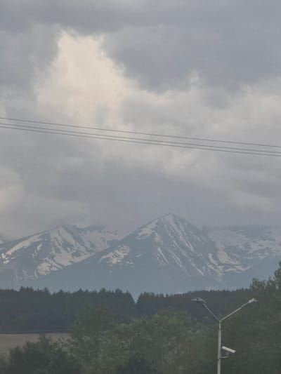 The image displays a vast, hazy mountainous landscape under an overcast sky. The scene is primarily composed of natural elements, with some man-made structures visible.
In the foreground, a dense, dark green forest stretches horizontally across the lower portion of the frame, marking a clear boundary. Above the forest, a majestic mountain range rises, its peaks and slopes partially obscured by a thick, grey haze or low-hanging clouds. Lighter patches, possibly snow or bare rock, are visible on the mountain's rugged surface, contrasting with its overall darker, subdued tone.
The sky, occupying the upper half of the image, is filled with varying shades of grey and white clouds, indicating an overcast day. Several thin, dark wires, likely power or telephone lines, traverse horizontally across the upper left section of the cloudy sky.
On the far right, extending from just above the treeline, is a utility pole structure. It features two angled arms, each equipped with what appears to be a light fixture or a camera, suggesting it might be a street light or surveillance equipment.
The weather appears hazy and diffused, contributing to a muted color palette dominated by greys, blues, and dark greens. The time of day is not distinctly clear but is daylight, likely under heavy cloud cover. There are no people visible, nor any specific activity taking place beyond the static natural environment. No text is discernible within the image. The location is Kraishte, Bulgaria.