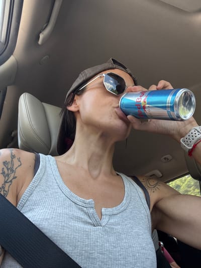 The image shows a woman in the driver's seat of a car, wearing a grey tank top, sunglasses, and a baseball cap. She is holding a can of Red Bull and drinking from it. She has tattoos on her left arm and shoulder. The car interior is visible, including the headrest and a seatbelt. The scene takes place in the city of Churek, Bulgaria.