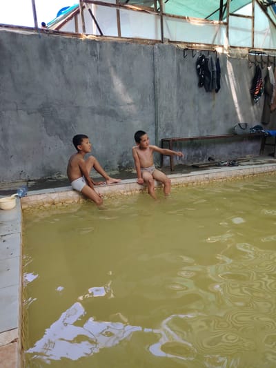 The image shows two young boys sitting on the edge of a pool, with their feet in the water. The pool appears to be outdoors, possibly in a rural or less developed area, given the concrete wall and the rustic overhead structure made of wood and a green tarp. The water in the pool is a murky, yellowish-brown color. On the wall behind the boys, clothing is hanging on hooks. A wooden bench is also visible, with a bag and some items of clothing on it. To the left of the pool, there is a plastic bottle and a small yellow bucket. The lighting suggests it is daytime, with sunlight filtering through the tarp and casting shadows on the wall. The boys appear relaxed, with one looking to the side and the other looking towards the water. The setting suggests a casual or recreational activity, likely a swim or a break in a hot climate. The location is identified as Kangli, Uzbekistan.