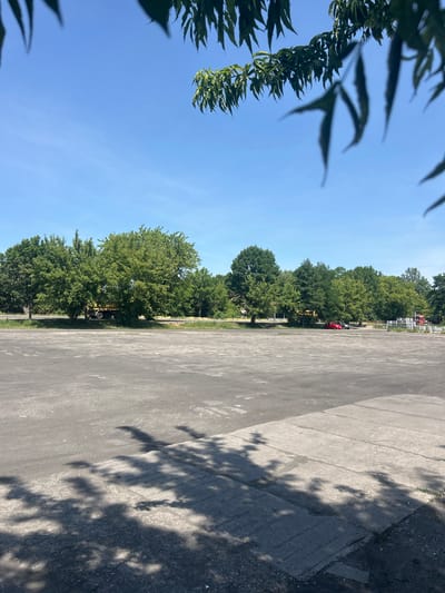 This image depicts an outdoor scene with a large asphalt lot in the foreground, marked by distinct shadows from overhead foliage. In the mid-ground, a line of mature green trees borders the lot. Behind the trees, a light yellow truck is partially visible, along with a red car and a dark-colored vehicle. Further to the right, stacks of grey materials and a red truck are present. The sky is a clear, bright blue. The location is identified as a parking area or open space in Konstancin-Jeziorna, Poland.