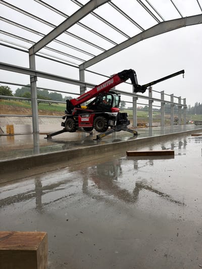An overhead shot captures the construction of a large building in Janowice, Poland, under a cloudy sky. The skeletal structure of the building, made of gray steel beams, dominates the frame. A red Manitou telehandler, equipped with a crane attachment, is positioned on the wet concrete floor. The telehandler's outriggers are extended for stability. The floor is covered in puddles, indicating recent rainfall. In the background, trees and a grassy slope are visible. A wooden beam rests on the concrete in the foreground.