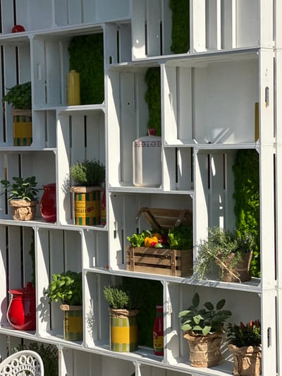 The image displays a white shelving unit constructed from wooden crates, filled with various plants and decorative items. The shelving unit is set against an outdoor backdrop, suggesting it is part of a garden or an outdoor market stall. The crates are arranged in a grid pattern, creating individual compartments.
In the top row, on the left, a red object, possibly an apple, rests on top of a crate. The second crate from the left contains green moss and a yellow ceramic vase. The third and fourth crates are empty.
The second row from the top features a plant in a yellow and green pot on the left, a red ceramic jar next to it, and another plant in a similar yellow and green pot. To the right, a white ceramic container with a red lid and decorative patterns is visible.
The third row contains a wooden crate filled with fresh vegetables, including lettuce, yellow bell peppers, and red bell peppers. To the right of this crate is a potted herb plant with green leaves, wrapped in burlap and tied with twine. Further to the right, there's another compartment with green moss.
In the bottom row, on the left, a red jar is placed next to a potted herb plant in a yellow and green container. To its right, another potted herb plant in a yellow and green container is visible. Further to the right, a bottle of ketchup is standing upright. On the far right, a potted plant with small red peppers is present.
The lighting suggests it is daytime, with sunlight casting shadows on the shelving unit and its contents. The overall impression is one of a rustic and decorative display, likely intended for a garden center, a farmer's market, or an outdoor cafe. The mention of Varenna, Italy, as the location implies this scene is situated in a picturesque Italian town, possibly along the shores of Lake Como, known for its charming streets and vibrant floral displays. However, there are no specific visual cues within the image that directly confirm this location.