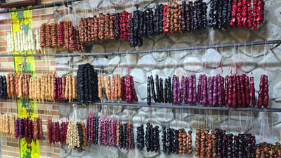 A shop in Manavi, Georgia, displays a large variety of churchkhela, a traditional Georgian candle-shaped candy made from grape must and nuts. The churchkhela are hung in rows on metal racks against a stone wall, with some colorful banners in the background. The candy comes in a wide range of colors, including white, brown, red, black, and purple, reflecting the different types of nuts and grape juices used in their preparation. The scene is well-lit, showcasing the texture and variety of the churchkhela. There are no people or animals visible in the image.