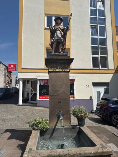 The image shows a street scene in Sankt Wendel, Germany, centered on a stone fountain with a statue of a man in historical dress holding a staff. The fountain is in the foreground, with water flowing from a spigot into the basin. In the background, a multi-story building with yellow and white facades and several windows is visible. A red sign for "PROFIL FAHRSCHULE" (driving school) is mounted on the side of a building to the left. A dark-colored car is parked in front of the building on the right. The street is paved with cobblestones, and there are planters with flowers near the fountain. The sky is clear and blue.