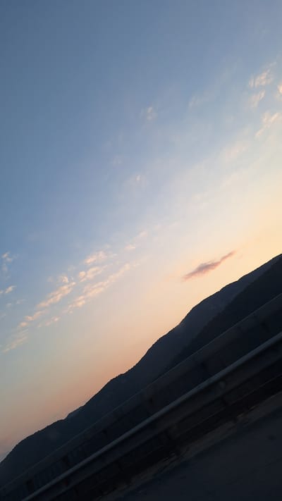 This image depicts a scenic view of a mountain range at sunset, likely taken from a road or elevated vantage point. The sky is a gradient of soft blues and pale oranges, with scattered wispy clouds tinged pink by the setting sun. A dark, silhouetted mountain range forms the horizon line. In the foreground, a metal guardrail runs diagonally across the frame, obscuring the lower portion of the scene. The overall impression is one of a peaceful twilight landscape. Without further information or context, the specific location within Vrachesh, Bulgaria, cannot be determined from the image alone, though it suggests a mountainous or hilly area within or near the town. There are no people or specific actions visible in the image.