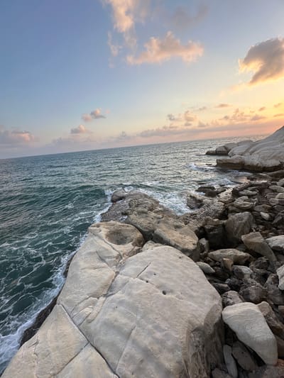 This is a landscape photograph taken at Rosh HaNikra in the Mate Asher Regional Council, Israel. The image captures a rocky coastline with the Mediterranean Sea in the foreground and a soft, colorful sky with scattered clouds in the background. The water is a deep teal color, with white foam from gentle waves lapping against the shore. Large, weathered limestone rocks dominate the right side of the frame, with smaller stones and pebbles scattered among them. The sky is a gradient of soft blues and pinks, transitioning into a warm orange hue towards the horizon, suggesting either sunrise or sunset. The lighting is diffused, creating a serene and natural atmosphere. No people or animals are visible in the image.