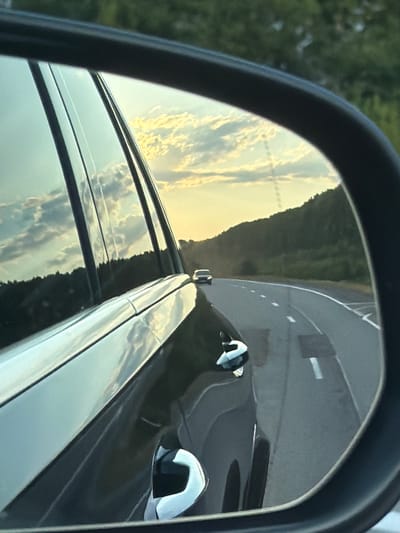 The image is a reflection in a car's side mirror, showing a road with a slight curve and a forested hillside. The sky is partly cloudy with the sun setting, casting a warm glow. Another car is visible on the road ahead. The reflection also shows part of the car the photo is being taken from, including a window and a door handle. There is no indication of Emel'yanovo, Russia in the image.