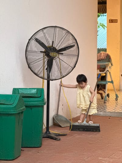 In Cổ Lũng Commune, Vietnam, a young child in a yellow dress is seen standing next to a large industrial fan. The child is holding a broom and appears to be sweeping the floor. To the left of the child are two large green trash cans. A dustpan is visible leaning against the wall behind the child. In the background, a high chair with a pink cloth is present. The floor is tiled with a patterned design, and the walls are a light color. The scene suggests an outdoor or semi-outdoor area, possibly a backyard or common space.