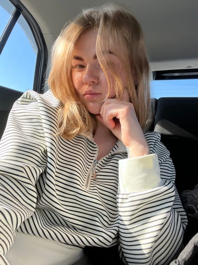 A woman with light blonde hair is reclining, possibly inside a car, as indicated by the window and car interior visible in the background. She is wearing a striped black and white zipped sweatshirt with white sleeves. Her hand is touching her cheek. The time of day appears to be daytime, evidenced by the bright blue sky outside the window. The location is not discernible from this image.