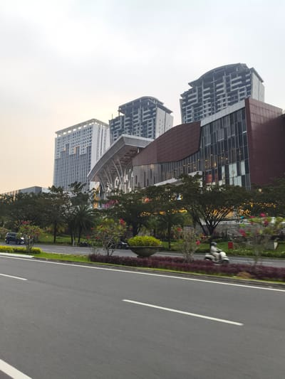 This image depicts a bustling urban scene in Babakan Madang, Indonesia, characterized by modern architecture, a multi-lane road, and lush greenery under an overcast sky. In the foreground, a wide asphalt road stretches forward, marked by distinct white lane lines. A green median strip separates the road, densely planted with mature trees, smaller shrubs, and flowering plants, including visible pink or red blooms and palm trees.
Several vehicles are present, indicating ongoing activity. A black car is partially visible on the left side of the median, while another dark vehicle is further down the road on the right. A white scooter, with a rider wearing a helmet, is captured in motion on the right lane, its blurred form conveying movement.
The background is dominated by a collection of tall, contemporary buildings. A prominent white high-rise with a distinctive grid pattern on its facade stands on the left. Adjacent to it is a large structure, possibly a mall or commercial complex, featuring a dark reddish-brown exterior with extensive glass paneling and a modern, undulating design. Behind these completed structures, several other high-rise buildings appear to be under construction or unfinished, showcasing their gray concrete frameworks against the horizon.
The time of day is difficult to pinpoint precisely due to the hazy or overcast sky, which casts a soft, diffused light over the scene. There's a faint warm, yellowish hue in the sky that could suggest either early morning or late afternoon, or simply a misty atmosphere. No discernible text is visible on any buildings, signs, or vehicles. The scene portrays an active, developing urban environment with a mix of finished and ongoing construction.