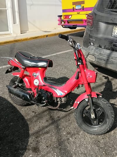 A red Honda Chaly moped is parked on an asphalt street in Puerto Plata, Dominican Republic. The moped is facing to the right and is partially obscured by a black SUV parked behind it. The SUV has a license plate that appears to be from the Dominican Republic. In the background, a yellow bus with purple trim is also visible. The street is lined with a yellow curb and a building with a white facade. The scene is illuminated by bright sunlight, casting shadows on the ground.