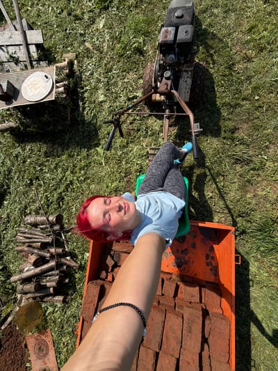 An overhead, high-angle shot captures a young woman with vibrant red hair lying on a green seat attached to an orange, rusted metal cart, which is itself connected to a red and black engine-powered tiller. The woman is reclined with her eyes closed and a relaxed expression, wearing a light blue t-shirt and grey patterned leggings. Her blue clogs are visible. The cart is filled with red bricks. To the left of the cart, a pile of cut logs lies on the grassy ground, next to a wooden structure and a circular metallic object. The scene is set outdoors in a grassy area, with shadows cast by the sun, indicating a daytime setting. The overall environment suggests a rural or suburban yard, possibly in Заболотово, Russia, during warmer weather.