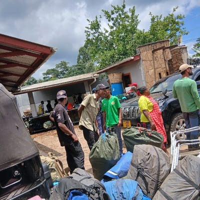 The scene depicts an outdoor staging area in Mweka, Tanzania, under a partly cloudy sky. Approximately twelve individuals, primarily adults, are present among vehicles and gear. In the immediate foreground, numerous large expedition-style bags, predominantly in shades of green, black, and blue, are stacked on the ground. To the far left, a dark-colored vehicle with a soft top is partially visible. In the center-left, three men are engaged in conversation; one wears a khaki shirt, another a green t-shirt with a yellow emblem, and the third a black t-shirt and baseball cap. To the right, a black Toyota Land Cruiser Prado SUV with a roof rack and Tanzanian license plate T 280 YEH is parked. A woman in a yellow top and red-patterned skirt/wrap stands near the SUV, alongside a man in a green long-sleeved shirt and a baseball cap. Metal folding chairs are visible near the rear of the SUV. In the background, several single-story buildings are present, including one with a corrugated metal roof and grey walls, and another featuring stone cladding. A blue barrel is visible near one of the buildings. A large, open-sided structure with a red-framed roof is situated on the far left. Lush green trees are visible above the structures, and the ground consists of packed earth.