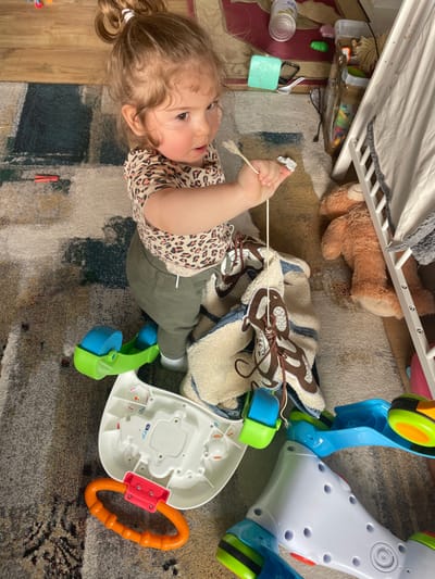 The image displays an indoor scene featuring a young child on a patterned carpet. The specific location in Senden, Germany, cannot be confirmed from the visual information provided.
A toddler, with light brown, curly hair secured in a high bun with a white elastic band, is visible in the center. The child wears a light-colored t-shirt with a brown leopard print pattern and olive-green pants, with white socks visible on the feet. The child is oriented slightly upwards and to the right, exhibiting an open-mouthed expression. The child's right hand grips a thin, light-colored stick-like object, possibly a skewer, alongside a small piece of white material. A thin string or cord descends from the stick, attaching to an off-white, textured blanket with brown, monkey-like patterns, which is partially draped over a toy.
The foreground and midground on the carpet contain several objects: an inverted white plastic toy base with attached blue and green components, another blue and green plastic toy piece, and an orange ring-shaped teether or handle with a red connector. To the right of the child, a brown teddy bear lies on its back adjacent to a white wooden structure, possibly a bed frame or doll's bed, partially covered by white fabric.
The background reveals a section of light brown wooden flooring and darker carpet, upon which various items are scattered, including a silver can, a light green plastic container, a white plastic fork, a red plastic clothespin, black-framed glasses, and a black cable. The floor throughout the visible area is covered by a multi-colored carpet featuring abstract patterns in shades of beige, grey, blue, and green. The scene is illuminated by ambient light.