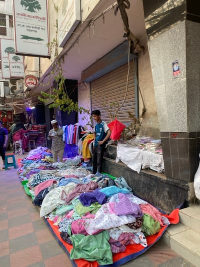 The image depicts a street market scene on S.K. Road, Narayanganj, Bangladesh, identified by signage for "RUPALI BANK PLC" and "ATM" with "NARAYANGANJ" visible.

In the foreground, a large quantity of assorted textile items, primarily clothing of various patterns and colors, is spread across a red and blue tarp on a tiled sidewalk. Two males are visible in the central frame. To the right, a young male, wearing a blue shirt with an England cricket logo and dark pants, stands with his hands on his hips, observing leftward. To his left, an older male, wearing a light-colored shirt and a white skullcap, stands behind the clothing display, appearing to hold a small, light-colored object in his hands.

The background features a building facade with a closed metallic roller shutter. Exposed wiring and roots or branches descend from an awning above the shutter. To the right of the shutter, a dark granite-like pillar shows a small framed picture adhered to its surface. Further left in the background, additional individuals are discernible, including one person in a patterned sari, suggesting ongoing commercial activity or pedestrian traffic. Several white signs with green logos, including "RUPALI BANK PLC" and "ATM", are mounted on the buildings overhead. A red plastic bag hangs near the younger vendor from a tree branch or wire. The ground is a tiled pedestrian walkway.