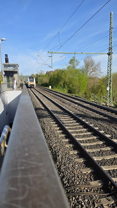 The image depicts a bright, sunny daytime scene at a railway station in Vellmar, Germany. The perspective looks along a set of train tracks that recede into the distance towards the upper left. The tracks are laid on dark ballast stones with concrete sleepers. Above the tracks, an extensive overhead line system (catenary) is visible, supported by green metallic gantries and numerous wires crisscrossing the clear blue sky.
On the right side of the image, a station platform runs parallel to the tracks. In the foreground, a light-colored concrete barrier or railing partially obscures the immediate trackside. Further along the platform, a modern-looking glass-enclosed shelter or small station building can be seen. Signs on this structure include the German word "Ausgang" (Exit), a wheelchair accessibility symbol, and separate male and female restroom symbols. Two small figures, presumably people, are visible near this shelter. A lamppost and a traffic light post also stand on the platform.
In the distance, on the leftmost track, a white and orange/yellow commuter or regional train is approaching or departing. The background on the left of the railway features a dense line of green trees and bushes, indicating a natural landscape alongside the tracks. The clear sky and strong shadows suggest bright sunlight. The overall scene captures the functional infrastructure of a modern railway line on a pleasant day.