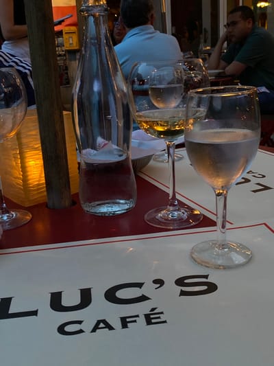 The image captures an outdoor dining scene at Luc's Cafe in Ridgefield, United States, during evening hours. In the foreground, a table displays a white placemat with the black text "LUC'S CAFÉ" on a red tabletop. Three stemmed glasses are visible: one empty on the far left, one containing a light amber liquid (wine) in the center, and one containing clear, cold liquid (water) on the right. A clear glass bottle, approximately half-full of water, stands centrally. A wooden post supports an illuminated, textured paper lantern emitting a warm, orange-yellow light on the left. In the midground, three individuals are partially visible at other tables: one with their back to the camera wearing a light blue shirt, a person with dark hair, and a third person in a dark green shirt with glasses, resting their chin on their hand. The background reveals an indoor portion of the cafe, a partial red French flag, and a yellow metal post box.