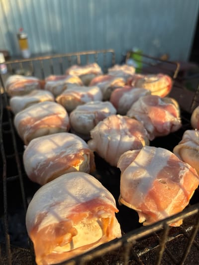 A close-up, eye-level shot shows a grill rack filled with appetizers wrapped in bacon. The appetizers appear to be small, round objects, possibly mushrooms or cheese balls, tightly encased in strips of raw bacon. The bacon is pale pink with white fat marbling, and some pieces have visible streaks of uncooked meat. The grill rack is dark metal, with a coarse grid pattern. The background is blurred, but suggests an outdoor setting with a light-colored wall and possibly some plants or foliage. The lighting indicates it is either early morning or late afternoon, with warm sunlight casting shadows. The scene appears to be set in a domestic backyard in the village of Dunilovo, Russia, given the context provided by the user.