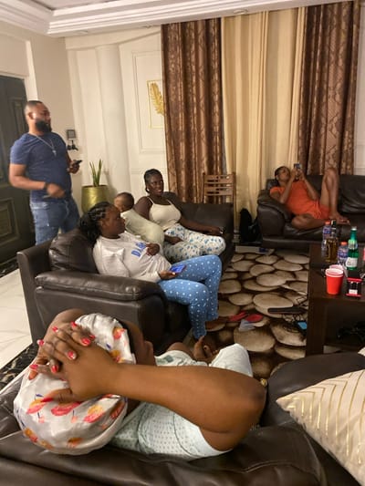 This image depicts a casual indoor gathering of approximately eight individuals, likely family or friends, in what appears to be a living room in Adenta Municipality, Ghana. The setting suggests an evening or nighttime occasion, indicated by the interior lighting.
Several people are relaxing on dark brown leather-like sofas. On the right side of the frame, one person in an orange outfit is engrossed in their mobile phone, while another person in blue polka-dotted pants also focuses on their device. A third individual on the sofa is wearing a light-colored sweatshirt, possibly with the text "DAB" visible, also looking at a phone. Further right, a person is seen with a floral silk bonnet covering their head, their hands clasped over it, revealing red nail polish.
Towards the center-left, a woman in a white top smiles towards the viewer, with a small child partially visible beside her. In the bottom left, a man wearing a blue t-shirt and jeans is partially visible, appearing to be standing or sitting, possibly also using a phone.
The room is furnished with a dark wooden coffee table, laden with several beverage bottles and red plastic cups. A textured rug with a pebble-like pattern covers the floor beneath the table. Brown patterned curtains are visible on the walls, alongside a gold-framed art piece, possibly featuring a feather design. Decorative pillows, including one with a gold geometric pattern, adorn the sofas. A potted plant adds a touch of greenery to the decor. The overall atmosphere is one of leisure and relaxation, with many individuals engaged with their mobile phones.