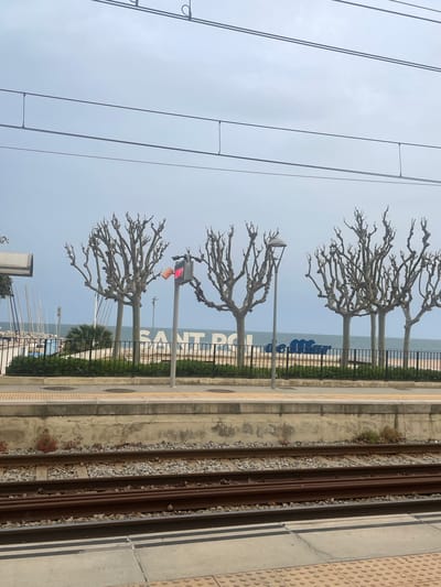 The image shows a view from a train platform in Sant Pol De Mar, Spain. The platform is on the right side of the frame, with textured, gray concrete slabs. To the left of the platform are train tracks, with visible gravel between the rails. Beyond the tracks, there's a concrete barrier followed by a decorative black metal fence. Trees with bare branches are planted along the fence line. In the background, the sea is visible with a large sign spelling out "SANT POL" in blue letters. A traffic light with a red light is positioned near the middle of the frame. Overhead electrical wires for the train run across the top left of the image against a pale blue sky. A few boats are visible in the water.