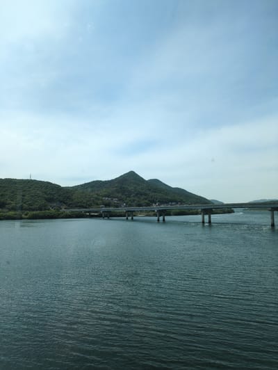 A panoramic landscape captured during the daytime features a range of lush, green mountains, a wide body of water, and a bridge under a light blue sky. The sky, occupying the upper portion of the image, is mostly clear with soft, diffused white clouds.
The left side of the frame is dominated by multiple mountain peaks, densely covered in vibrant green foliage. Small structures or buildings are faintly visible nestled within the green slopes, and a tall, slender transmission tower or mast stands on a lower mountain towards the far left.
To the right, a significant expanse of water, possibly a river or lake, stretches horizontally. The water's surface is a deep teal or bluish-green, characterized by numerous small ripples that indicate gentle movement or a light breeze.
A long, multi-span bridge traverses the body of water, running parallel to the mountain range. It is supported by several robust concrete pillars that extend into the water. The scene suggests a tranquil setting in Yangsan-si, South Korea, with no people or visible text. The bright, even lighting indicates clear weather, likely in the late morning or early afternoon.
