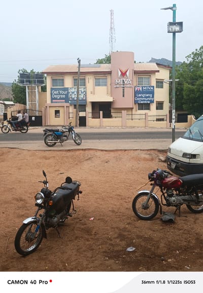 The image is taken in Langtang, Nigeria, with several motorcycles prominently featured. In the foreground, two motorcycles are parked on a reddish-brown, dusty ground. One motorcycle on the left is black, and the other on the right is red with chrome accents.
In the background, across a paved road, a two-story building is visible. The building is painted in beige and brown hues. The building bears the logo of "MIVA Open University" and a sign for "Langtang Community Centre." A banner advertising "Get Your Degree" is also attached to the building. A water tower and a communication tower are visible behind the building.
On the road, a blue motorcycle is parked, and another motorcycle with two people on it is in motion. A white van is parked on the right side of the road. A green lamp post stands near the van.
The overall environment appears to be urban with a mix of residential and commercial buildings. The sky is overcast, suggesting diffused lighting.
