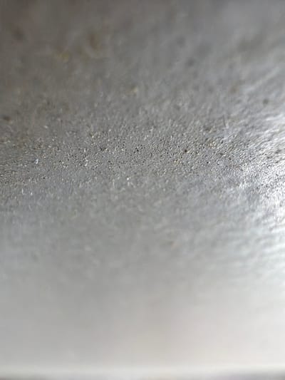 The image is a close-up, abstract shot of a textured, greyish surface. The texture appears granular, with tiny dark specks embedded in a lighter matrix. There are some areas that seem slightly raised or bubbled, and a diffuse sheen across the surface, suggesting it may be slightly damp or have a glossy finish. The lighting is soft and diffused, creating a shallow depth of field with the foreground in sharper focus and the background becoming progressively blurred. The overall impression is one of a very close view of a material like concrete, textured paint, or possibly even a dried liquid with particulate matter. There are no discernible people, objects, actions, or interactions. The location in Grapevine, United States of America, cannot be determined from the visual information presented in the image.