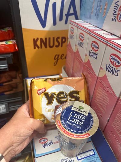The image shows a hand holding a package of "yes" caramel chocolate bars. In the background, there are several boxes of "Dr. Oetker Vit" cereal and "Alnatura Caffè Latte Macchiato" cups. The overall scene suggests a retail environment, possibly a supermarket or grocery store, with products displayed on shelves. The presence of German text on the packaging indicates the location is likely in Germany, and given the city of Tuntenhausen, Germany, this is a plausible setting for such a display. There are no specific details about the environment beyond the product displays. The lighting is consistent with indoor retail lighting.