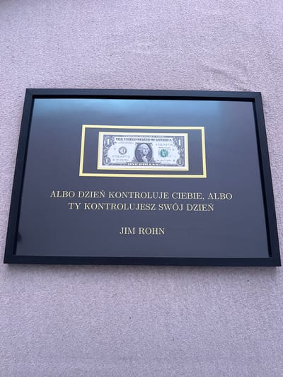 The image shows a framed one-dollar bill with a quote below it. The quote is in Polish and reads: "ALBO DZIEŃ KONTROLUJE CIEBIE, ALBO TY KONTROLUJESZ SWÓJ DZIEŃ" which translates to "Either the day controls you, or you control your day." Below the quote is the name "JIM ROHN". The framed item is displayed on a textured gray surface, possibly a rug or fabric. The location in the city of Kobylnica, Poland is not discernible from the image.