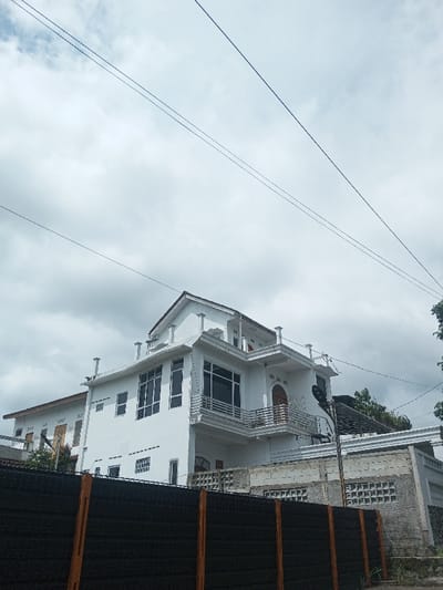 A modern, white two-story building stands under a cloudy sky, its design featuring large windows and a balcony with metal railings. A dark fence with orange posts obscures the lower portion of the building and a section of a gray concrete wall with decorative openings. Power lines crisscross the sky above, adding a geometric contrast to the natural cloudscape. Trees peek out from behind the building, suggesting a suburban or semi-rural setting.