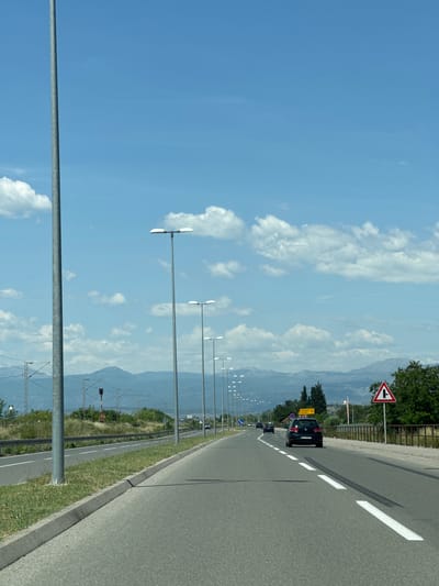 The image depicts a road running through Goričani, Montenegro, with mountains visible in the background. Several lampposts line the road. A car is present on the road, along with a road sign indicating a junction. The sky is blue with scattered clouds. Overhead power lines and supporting structures are also visible. The road has painted lane markings.
