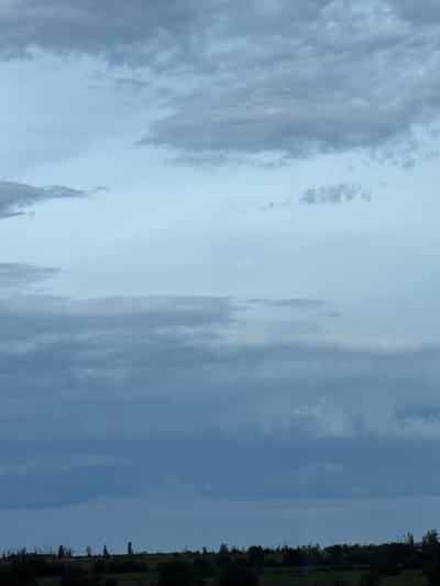 The image depicts a landscape scene with a dark, overcast sky dominating the upper portion. The lower part of the image features a silhouetted treeline, suggesting a rural or natural environment. The sky is filled with layered clouds of varying shades of gray, indicating a potentially moody or approaching storm. The absence of specific landmarks or human activity makes it difficult to pinpoint an exact location within Gelemenovo, Bulgaria, but the general scene is one of an open, natural space under a heavily clouded sky.