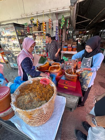 In Grong-Grong, Indonesia, a market scene unfolds as two women and a man engage in transactions. A woman wearing a pink hijab serves customers from behind a stall laden with food items. She is positioned next to a large basket overflowing with dried noodles, a staple in Indonesian cuisine. Beside her, another woman, also in a hijab, is actively preparing food, scooping a spicy dish from a large container into a plastic bag. Her apron, adorned with an abstract design, suggests a vendor's uniform. A man, dressed in a plaid shirt and a black cap, stands nearby, observing the interactions.

The stall itself is a testament to the vibrant street food culture. Various containers, some plastic and others glass, hold an array of colorful ingredients and prepared dishes, hinting at the diverse culinary offerings. The background reveals a bustling market environment with shelves stocked with goods, suggesting a lively commerce. Overhead, strings of packaged snacks hang, adding to the visual texture of the scene. The ground is a mixture of dirt and pavement, typical of an outdoor market. The overall atmosphere is one of everyday commerce, capturing a slice of life in Grong-Grong.