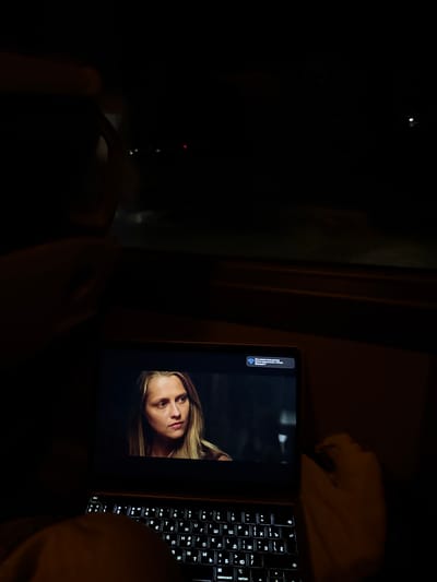 Here is a forensic description of the image:
The image shows a laptop open in a dimly lit environment. A person is visible on the screen, appearing to be an actress in a movie or show. The keyboard of the laptop is illuminated, suggesting it is in use in a low-light setting. To the right of the laptop, a blurry object suggests a person's arm. The background is mostly dark, with a window and possibly some indistinct shapes. There's no indication of the location within Cajetina Municipality, Serbia. There is no other information to give that allows for a description of objects or people.