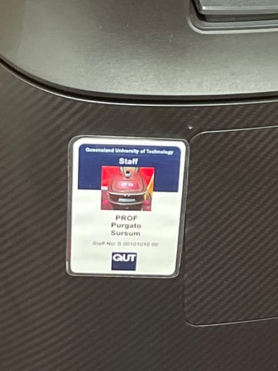 The image features an identification card from Queensland University of Technology (QUT) resting on a dark gray surface, likely a laptop or similar device, characterized by a fine diagonal textured pattern resembling carbon fiber. The ID card is for "Staff" and displays the name "PROF Purgato Sursum" along with a staff number: "S 00101010 00". A small, somewhat blurry photograph of a person is visible on the card, and the QUT logo is present in the bottom right corner. The card is enclosed in a clear plastic holder. No specific activity is taking place, and no individuals are visible. The setting appears to be indoors, possibly an office or desk environment, consistent with a university context in Brisbane City, Australia.