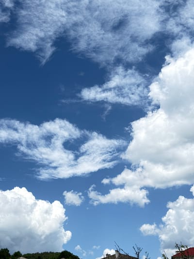 The image depicts a view of the sky in Ogradna, Bulgaria. Predominantly blue, the sky is partially covered by various types of clouds, including cirrus and cumulus. The clouds range from thin, wispy formations to larger, puffy masses. Fragments of buildings and trees are visible along the right edge of the frame.