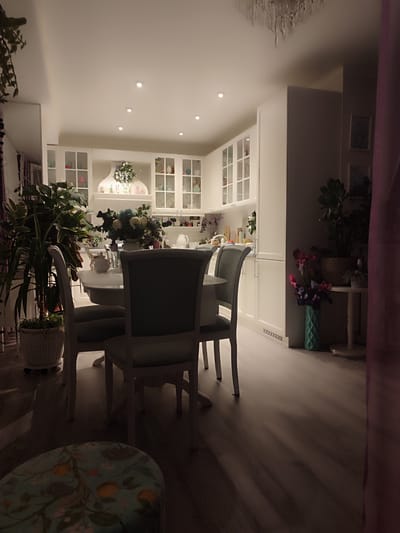 This is an interior scene, likely a dining area and kitchen within a home. The location is unknown. The space is lit by ceiling lights, casting shadows across the wooden floor. The foreground features a fabric-covered ottoman with floral patterns. A round white table is surrounded by three dining chairs with light-colored frames and upholstered seats and backs in a muted color. On the table sits a floral arrangement in a white vase, along with other tableware. A potted plant in a woven basket stands beside the table.
In the background, white kitchen cabinets with glass doors line the wall. A display shelf above the countertop holds decorative items. Another potted plant is visible near the cabinets. To the right, a side table holds another plant and a vase with flowers. A partial view of a curtain in the right foreground adds depth.