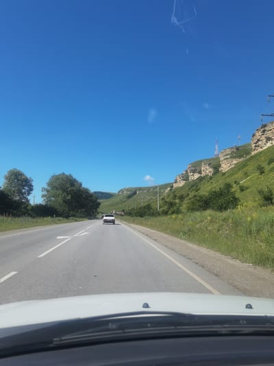 The image captures a view from inside a vehicle traveling on a paved road in Khabez, Russia. The road has visible white lane markings, including a directional arrow indicating a turn or split in the road. A white car is driving ahead in the same lane. The landscape features a steep, grassy hillside with rocky outcrops on the right side of the road, dotted with green trees and vegetation. Power lines and a tall transmission tower are visible on the hillside. The sky is clear and bright blue. On the left side of the road, there are several large green trees and a grassy verge. The foreground shows the dashboard and windshield of the vehicle.