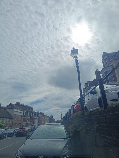 The image depicts a street scene with parked cars and buildings in Arun, United Kingdom. The sky is cloudy with the sun partially visible, creating a bright spot. A vintage street lamp stands on the right side of the frame, illuminating the path. Several cars are parked along the street, including a white car and an orange car. The buildings on the right are made of brick and have traditional architectural features. The street is paved, and there are brick walls lining the side of the road. The overall impression is a quiet, residential street in an English town.