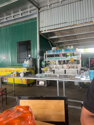 The image shows an outdoor area of a food establishment, likely a beer garden or restaurant, located in Hợp Châu, Vietnam. The scene is characterized by its utilitarian construction, with corrugated metal walls and roofing, and a generally open-air feel.

In the foreground, a table is set with glasses and a small teapot, suggesting immediate use or recent service. Partially obscuring the table is an out-of-focus plastic bag containing orange-colored items.

The middle ground is dominated by a stainless steel bar or counter area. On this counter, there are various items, including a beer dispenser, stacks of blue cups, numerous glasses on shelves, and a sink. A large, illuminated display featuring a beer advertisement is visible behind the counter.

The background reveals more of the structure, with visible metal beams supporting the roof and a green corrugated metal wall on the left. The overall impression is of a functional, perhaps slightly rustic, establishment catering to outdoor dining and drinking. The environment appears to be a commercial zone within Hợp Châu, given the presence of a business and its associated amenities. There are no visible people interacting in the scene.