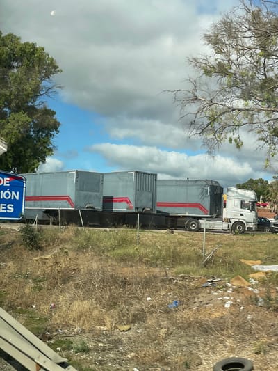 This is a vertically oriented, slightly tilted, eye-level medium shot of several large, gray shipping containers with red stripes, some of which are stacked. They are parked side-by-side on a paved surface, next to a ditch filled with dry grass, weeds, and scattered litter. A white semi-truck cab is attached to the front of the first container. In the background, trees and a cloudy sky are visible. The scene appears to be outdoors during the daytime, with overcast conditions.

To the left of the containers, a tree with bare branches extends into the frame, partially obscuring the sky. A blue sign with white lettering is visible at the bottom of the frame, in front of the containers. The text on the sign is partially obscured but appears to read "DE" and "ION ES".

The overall impression is one of industrial storage or transportation, possibly in a somewhat neglected or utilitarian area, given the presence of litter. The lighting suggests it is daytime, but the clouds indicate potentially mixed weather. The location context of Los Barrios, Spain, suggests a possibly Mediterranean climate. There are no people clearly visible in the image.