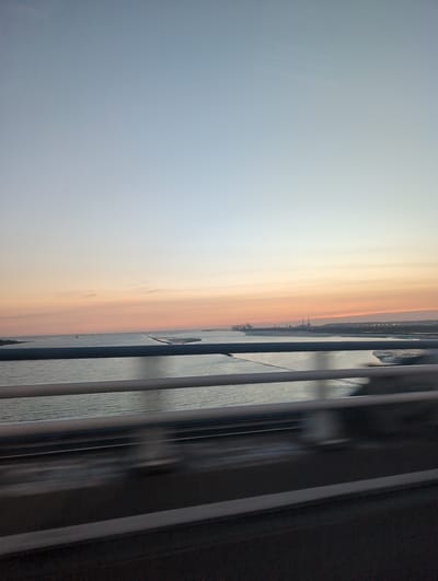 The image captures a serene sunset over a body of water, possibly a bay or a river, with a silhouetted industrial landscape in the distance. The sky transitions from a pale blue at the top to warm hues of orange and pink closer to the horizon, indicating the setting sun. Several ships are visible on the water. In the foreground, there are railings, suggesting the photo was taken from a bridge or a similar elevated structure. The water is calm, with gentle ripples reflecting the sky's colors. The industrial silhouette includes numerous structures, likely factories and port facilities, characteristic of a coastal or riverside industrial zone. The overall atmosphere is peaceful and tranquil, enhanced by the soft light of dusk. The location is identified as Sandouville, France, known for its port and industrial activities.