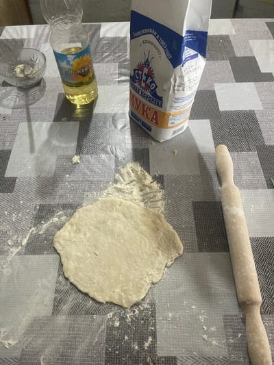 The image shows a table covered with a patterned tablecloth, where dough is being prepared. Scattered across the table are several objects related to baking. In the upper right corner, a wooden rolling pin lies horizontally. To the left of the rolling pin, a flattened disc of dough is visible, dusted with flour. Next to the dough, a bag of flour is partially visible, with text in Russian, including "ХЛЕБОПРОДУКТ-1" and "Мука". In the lower left corner, a partially empty plastic bottle of sunflower oil with a label featuring a sunflower and Russian text "Нежинская" is placed horizontally, next to a small glass with some liquid. The overall scene suggests the preparation of some baked goods, likely bread or pastries, in a domestic setting. The lighting is moderate, suggesting an indoor environment, possibly during the daytime. The background tablecloth has a geometric pattern of squares and zigzags in shades of grey and white. There are no people visible in the image, and no clear indication of the time of day or specific weather conditions.