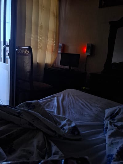 The image depicts a bedroom interior in Armavir, Armenia, at dawn or dusk. The scene is dimly lit, with light filtering through sheer vertical blinds on the left, revealing a glimpse of an exterior balcony with a railing. In the foreground, a bed is covered with crumpled white and floral-patterned linens. To the left of the bed, a dark wooden chair with a woven back is partially visible. In the background, a dark wooden dresser or cabinet is situated against a wall. A computer monitor rests on the cabinet, and to its right, a small speaker emits a red light. Another speaker is visible to the left of the monitor. The overall atmosphere is quiet and still, suggesting a private residential space.