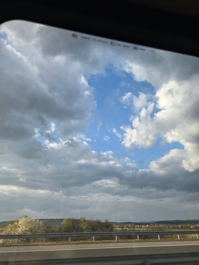 The image captures a rural landscape viewed from a moving vehicle, evidenced by the curved window frame and reflections. The sky is largely overcast with dark grey stratocumulus clouds, yet a prominent, irregularly shaped patch of clear blue sky with white cumulus clouds is visible in the upper-center. The lower third of the frame shows a multi-lane asphalt road with a white demarcation line, bordered by a two-tier metal guardrail on the right. Beyond the guardrail, the terrain consists of rolling hills and fields with interspersed vegetation. Some deciduous trees with white blossoms, indicative of spring, are present in the midground, alongside other trees with budding green foliage. In the far background, the horizon features faint outlines of distant hills and what appear to be scattered structures. No individuals are visible. Faint reflections, resembling a digital display's status bar elements (e.g., signal strength, battery icon), are present on the upper window surface. The scene depicts a highway or major road passing through an agricultural or semi-natural area. No discernible landmarks confirm the specific location as Yazdach, Bulgaria; the landscape is generic to rural regions.
