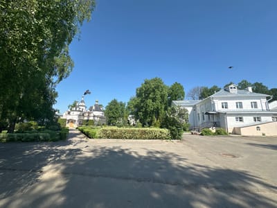 The image depicts a view of a street leading to a building, likely a church or monastery, in Махра, Russia. A paved road with tree shadows covers the foreground, leading to a manicured lawn and a white building with domes, indicative of Russian Orthodox architecture. To the right, a two-story white building stands with windows and a staircase. Trees are scattered throughout the scene, contributing to a serene environment. A bird is visible in the sky, adding a sense of movement to the still composition.
