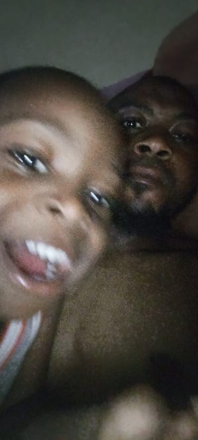 The image is a close-up, out-of-focus selfie taken in a dimly lit indoor environment, possibly a bedroom, in Rijiyar Zaki, Nigeria. Two individuals are visible: a child in the foreground, whose face is large and fills much of the left side of the frame, and an adult behind them, whose face is more in the background and partially obscured. The child appears to be sticking their tongue out. The adult's eyes and nose are visible, with a beard present. The background is dark and indistinct, suggesting low lighting conditions. The overall impression is of a casual, personal moment captured by the individuals themselves.